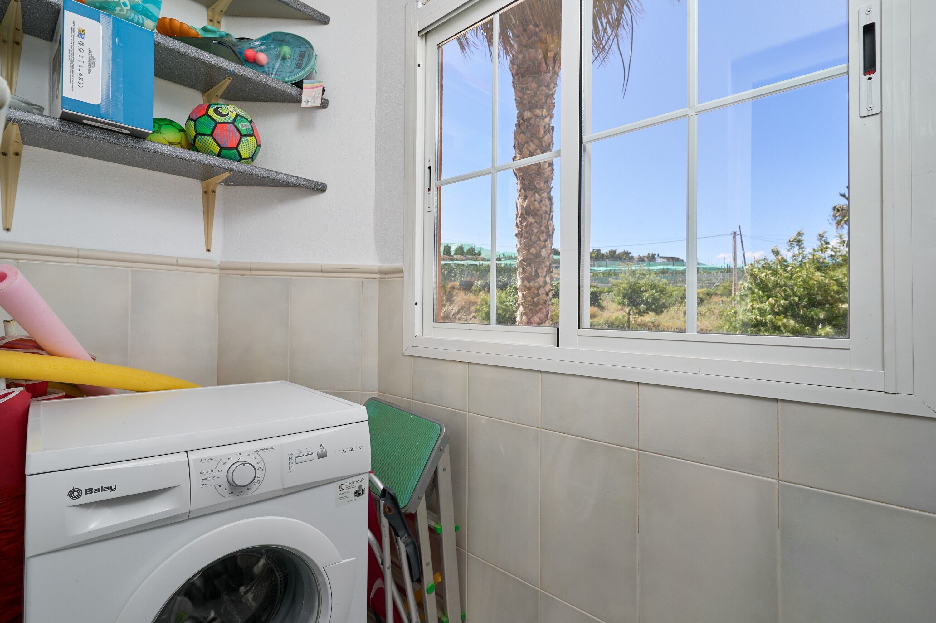 A laundry area with a washing machine, shelves with items, and a window with a view of greenery.