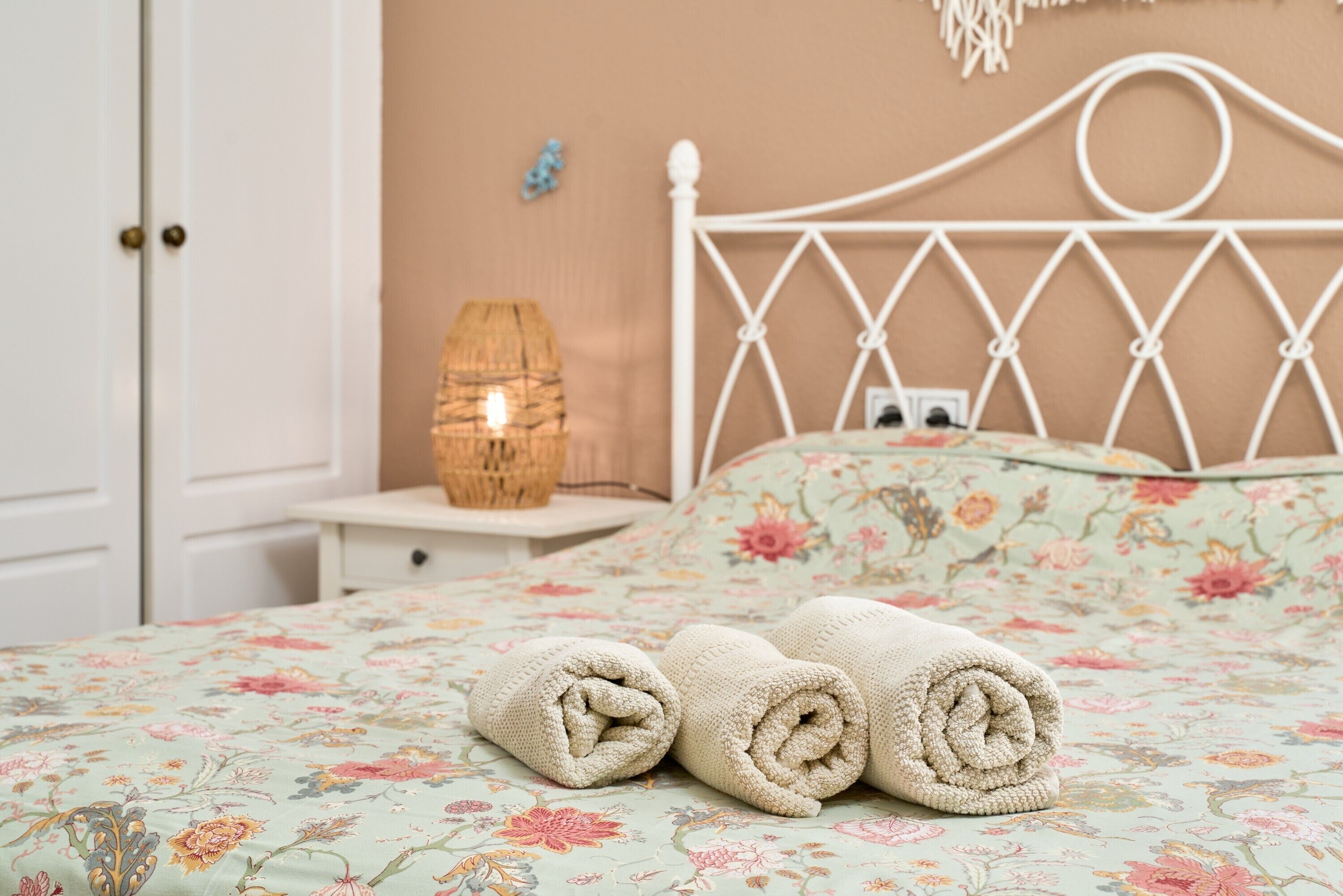 Ein ordentlich bezogenes Bett mit Blumenmuster, ein weißes Metall-Kopfteil und eine Nachttischlampe.