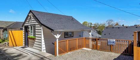 A small, single-story building with a sloped roof, a wooden fence, and a gravel driveway.