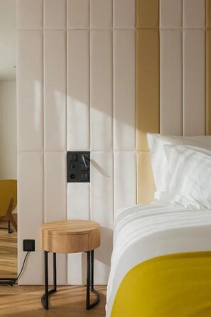 Une chambre d’hôtel avec un lit, un tabouret en bois, un panneau de commande et une chaise jaune.