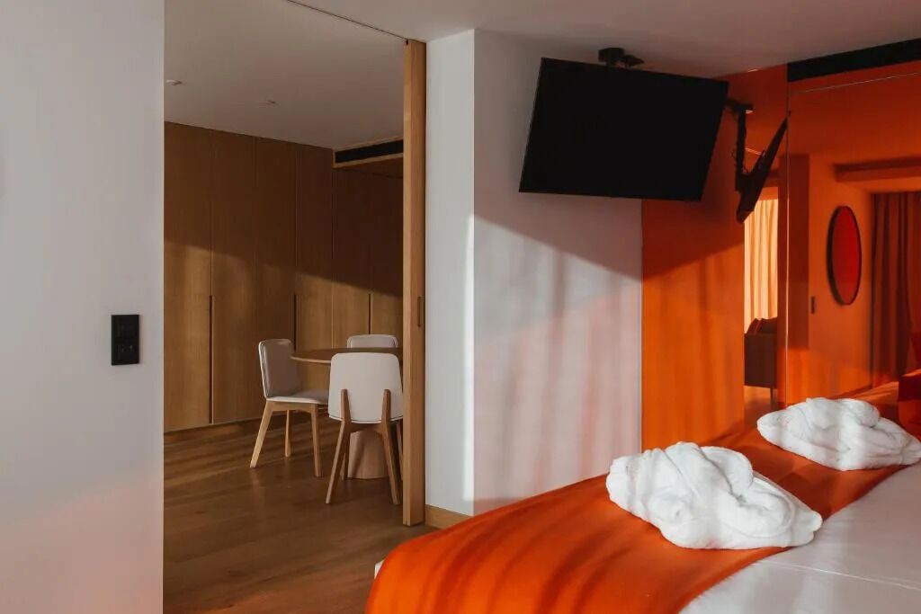 A hotel room with a bed, a TV mounted on the wall, a desk with a chair, and a wooden wardrobe.