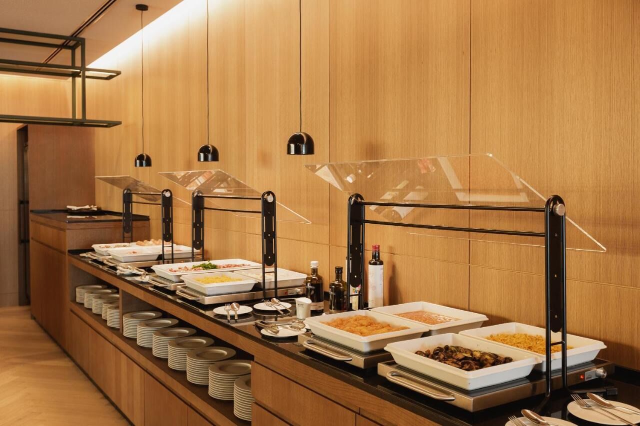 A buffet setup with clear plastic covers over food dishes, arranged on a long counter with stacked plates and condiments.