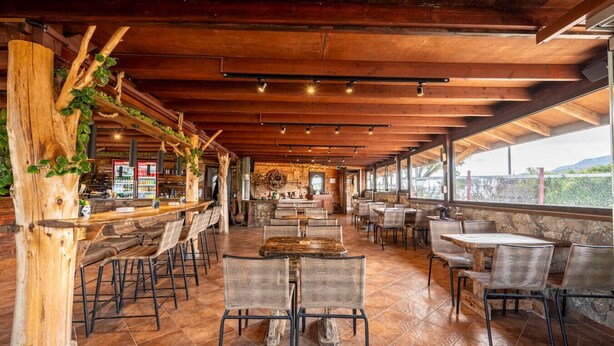 Ein Restaurant mit Holztischen und -stühlen, einer Bar und Blick ins Freie.