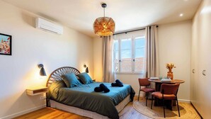 Quarto com cama, mesa de cabeceira, luminária, janela e um pequeno espaço de refeições com cadeiras e mesa.