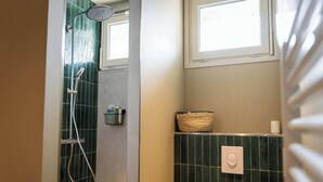 Uma casa de banho com chuveiro, porta-toalhas, cesto e suporte para papel higiénico.
