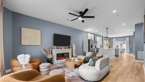 Ein modernes Wohnzimmer mit Kamin, Deckenventilator und sichtbarer Küchenzeile im Hintergrund.