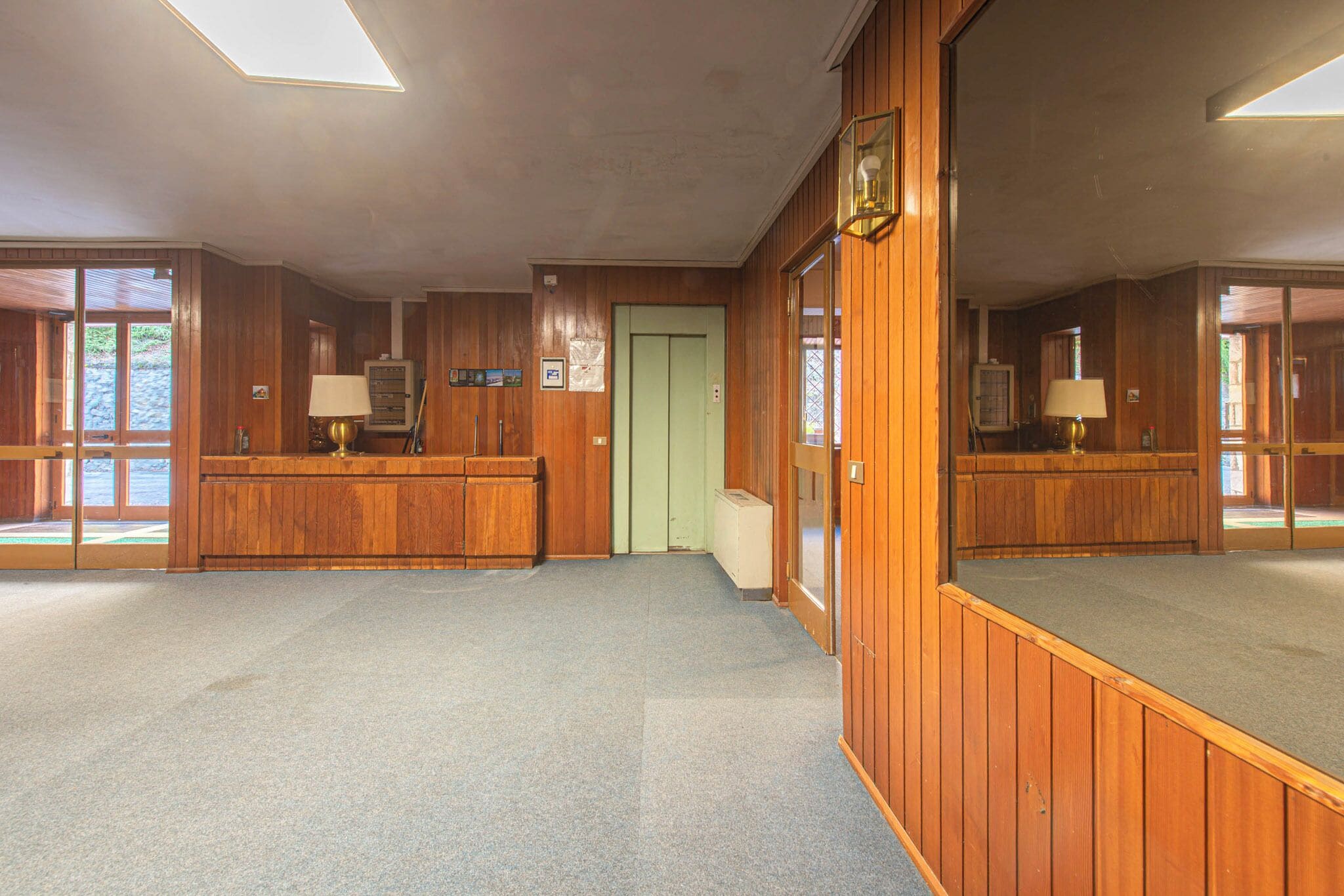 Vestíbulo de hotel con paneles de madera, una recepción, una puerta verde y un espejo grande que refleja la habitación.