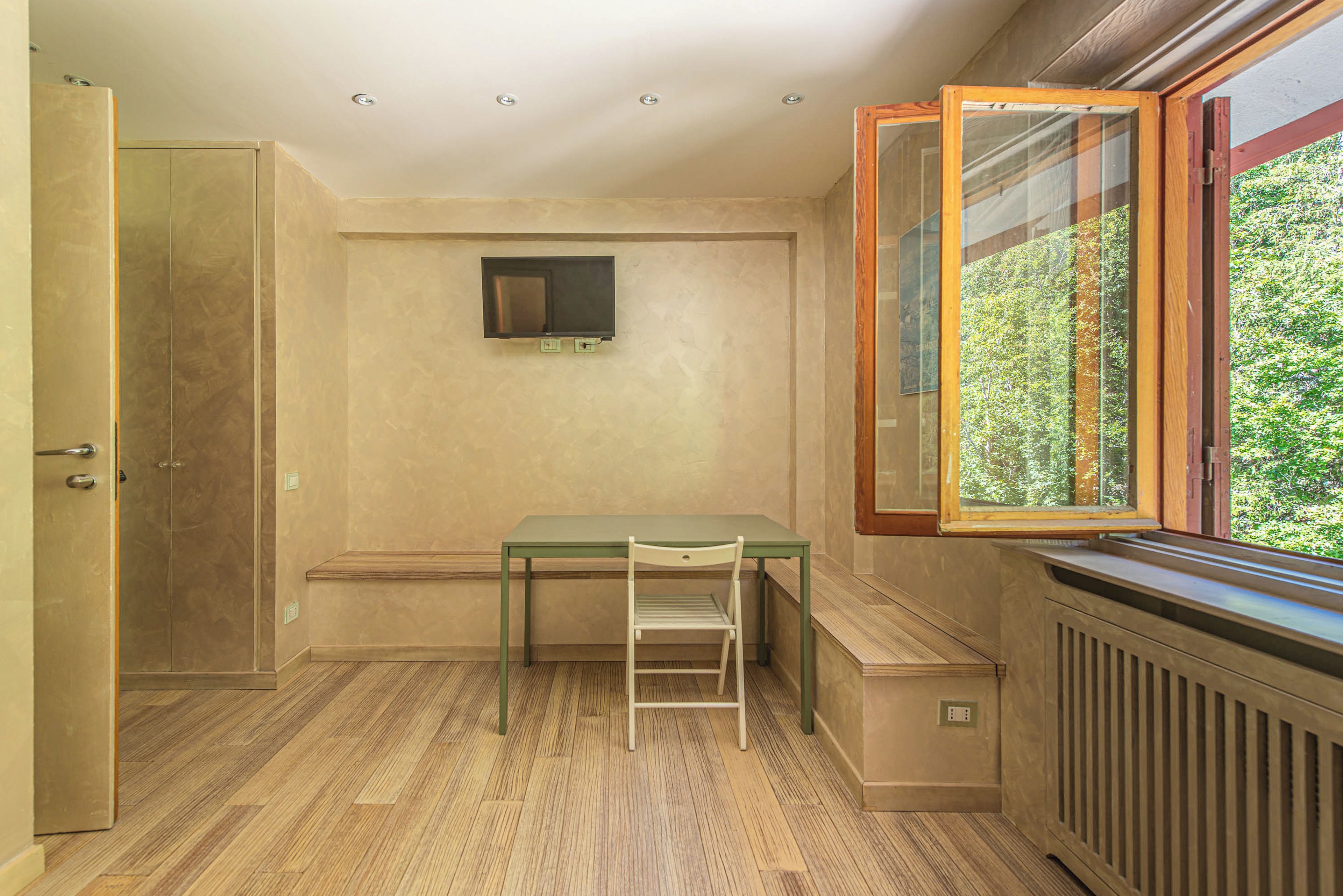 Una habitación con piso de madera, una mesa verde con una silla, un televisor montado, una ventana con vista a follaje y un radiador.