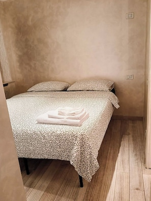Un lit simple avec une couvre-lit à motifs et deux oreillers, une serviette pliée posée sur le lit, et un plancher en bois.
