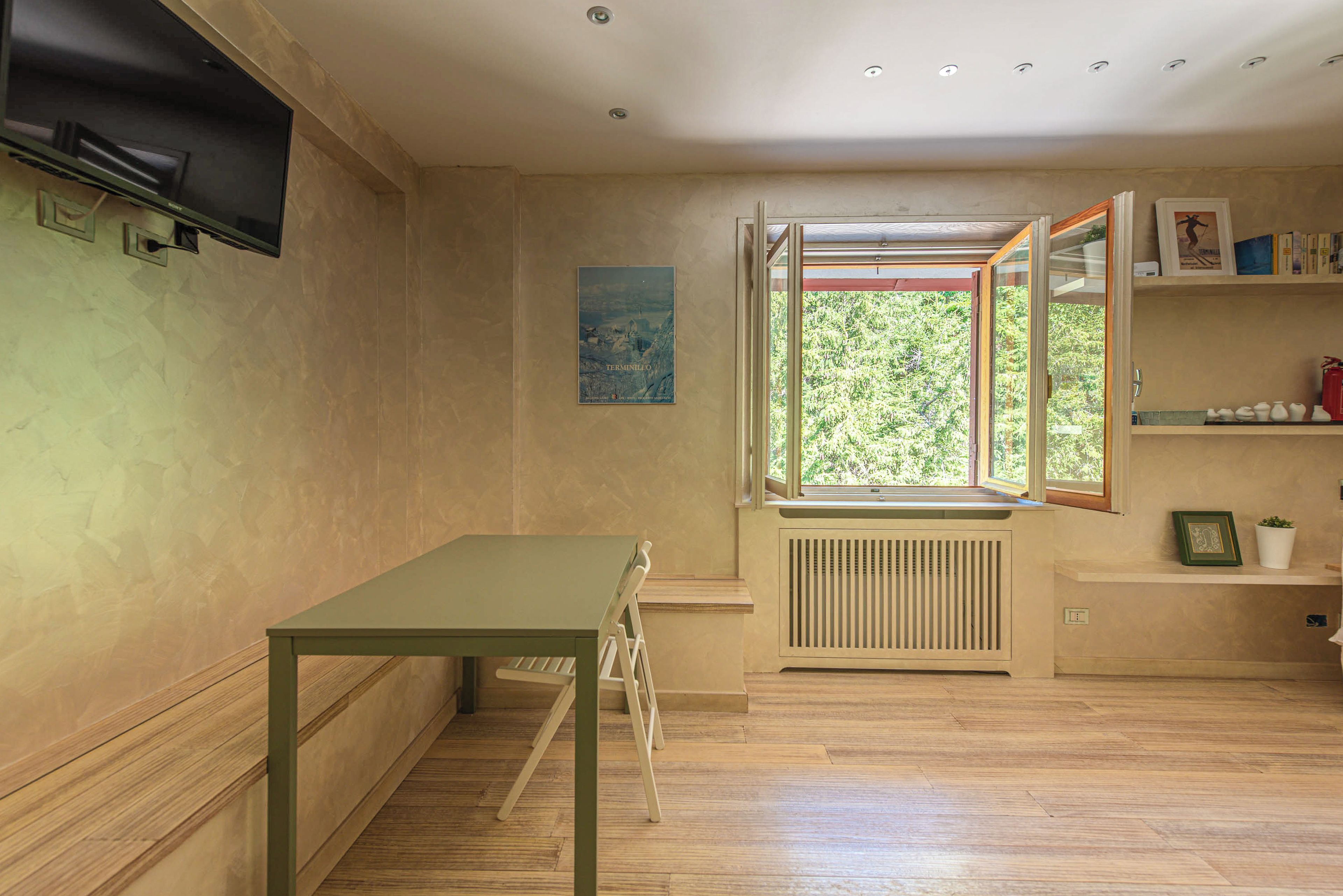 A room with a wooden floor, a table, a radiator, a window with an open view, and a shelf with books and decorative items.