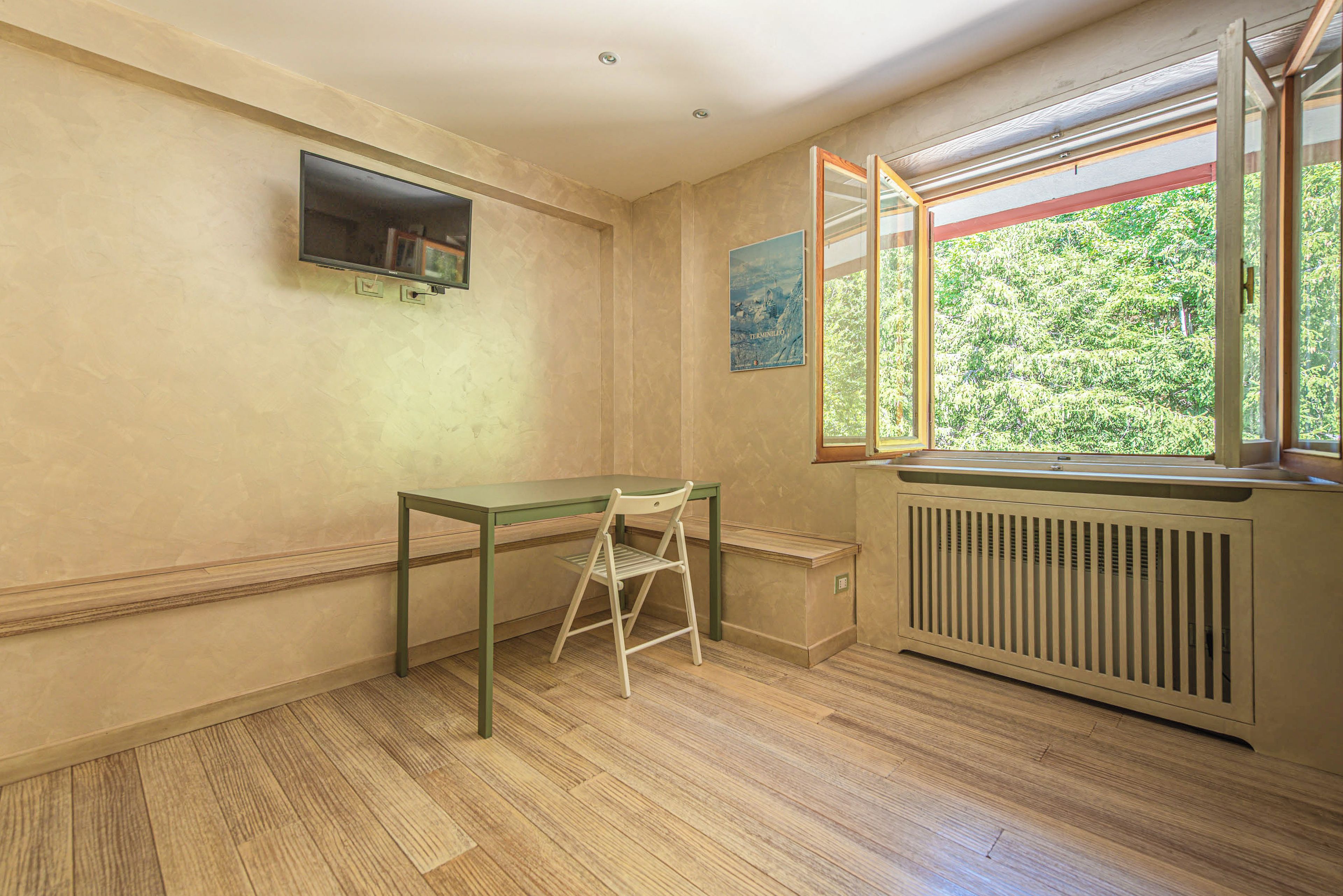 Una habitación con piso de madera, una mesa con una silla, un radiador y una ventana con vista a la vegetación.