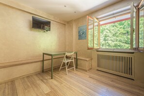 A room with a wooden floor, a table with a chair, a radiator, and a window with a view of greenery. - La Casina (Pian De' Valli)