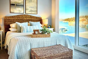 A bedroom with a large bed, a wicker headboard, and a view of a body of water through a large window. - Lake Views, Resort Perks! Osage Beach Retreat (Osage Beach)