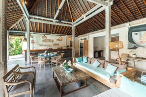 A spacious indoor area with a high ceiling, wooden beams, a seating area with cushions, a dining table with chairs, and a kitchen area in the background. - Stylish 4BR Canggu Villa • Private Pool & Sundeck • 8 Min to Beach (Badung Regency)