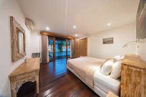 A bedroom with a wooden floor, a bed with white bedding, a wooden nightstand, a mirror, and a sliding glass door leading to a balcony. - Stylish 4BR Canggu Villa • Private Pool & Sundeck • 8 Min to Beach (Badung Regency)