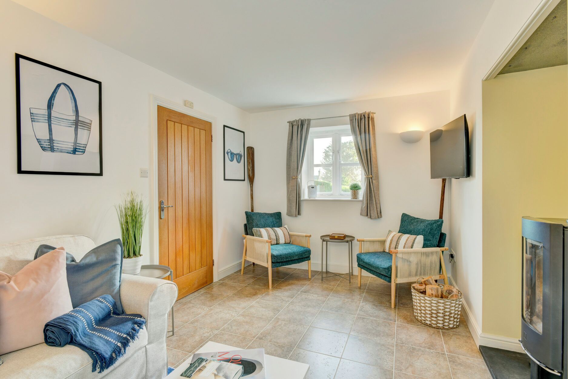 Un salon comprenant un canapé, deux fauteuils, une table basse et un téléviseur. On y trouve une porte, une fenêtre avec des rideaux et un téléviseur fixé au mur.