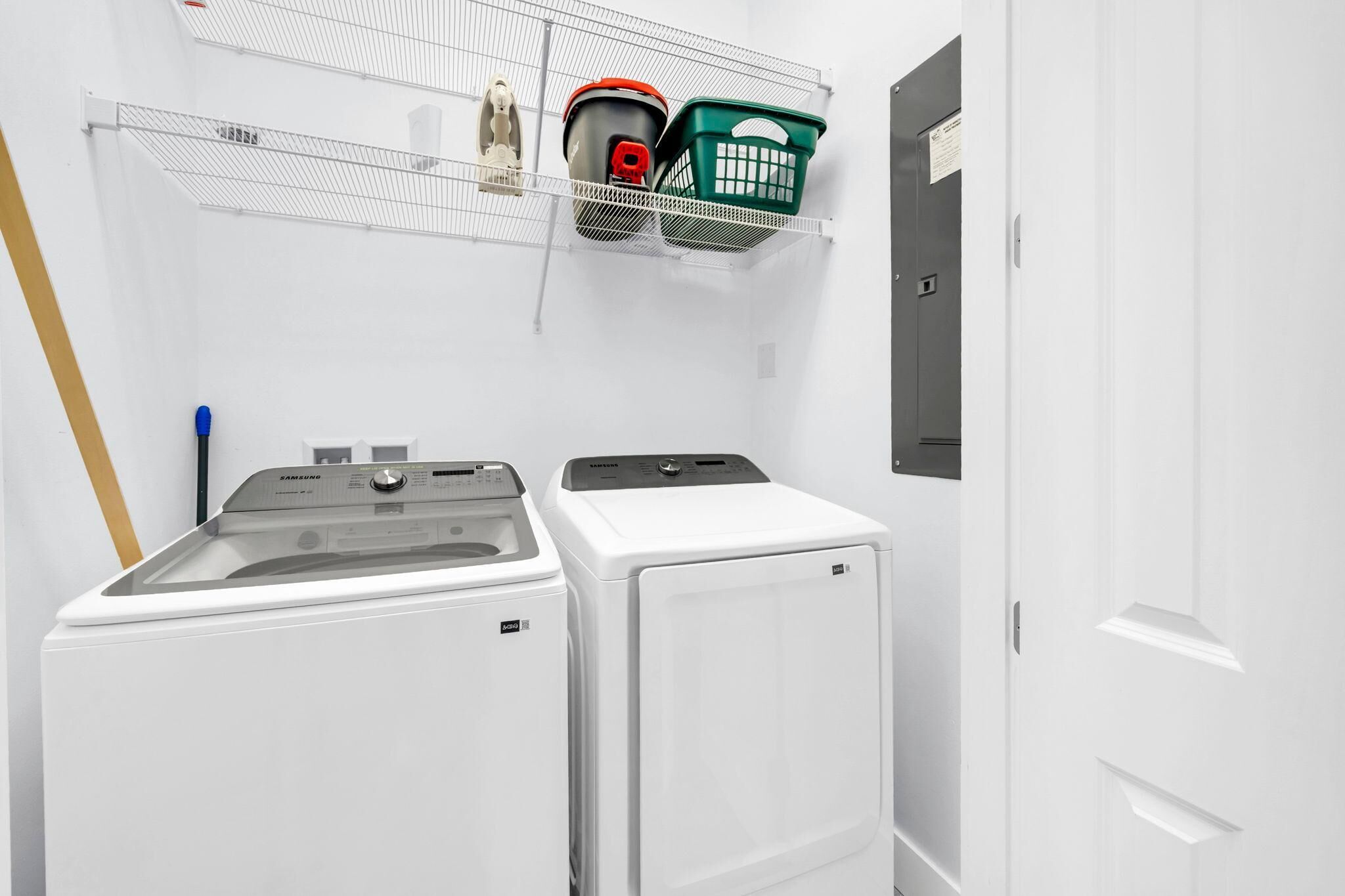 A laundry room with two white washing machines, a metal shelf with baskets, and a door with a sign.