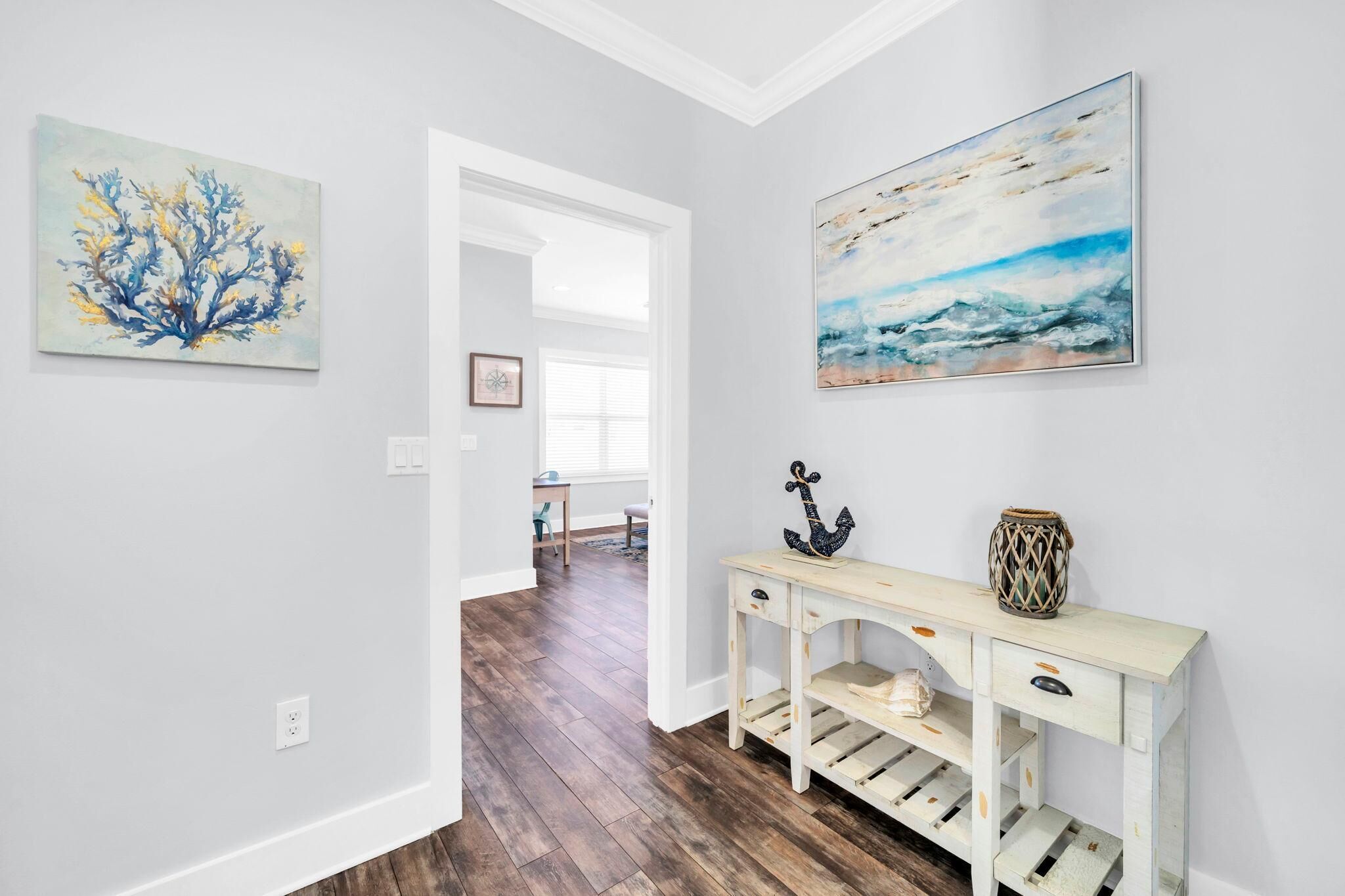 A hallway with a wooden floor, a white console table with decorative items, and two paintings on the walls.