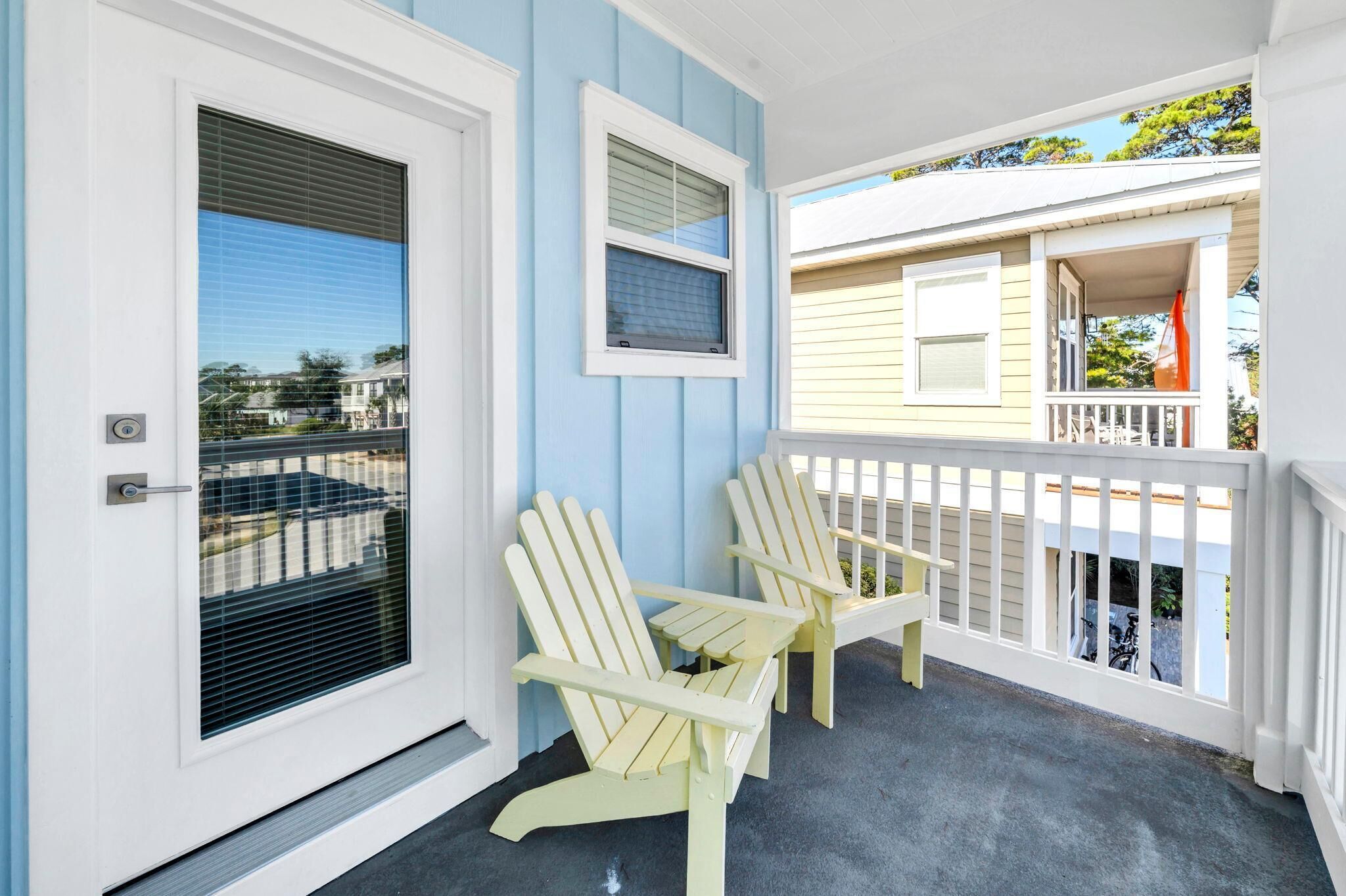 A porch with two Adirondack chairs, a white door with a window, and a balcony with a railing.