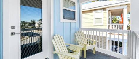 A porch with two Adirondack chairs, a white door with a window, and a balcony with a railing.