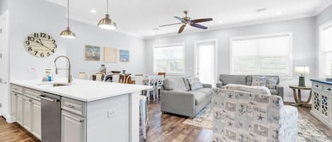 A modern kitchen with a central island, a dining area with a white table and chairs, a grey sofa, and a ceiling fan.