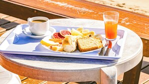 Un plateau de petit-déjeuner avec des toasts, des fruits et un verre de jus d’orange, posé sur une table près de la plage.