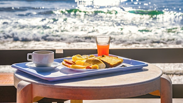 Un plateau contenant des aliments pour le petit-déjeuner, notamment des toasts, des fruits et une boisson, est posé sur une table au bord de l’eau.