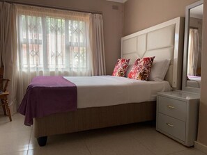 Un dormitorio con cama, mesita de noche, espejo y ventana con cortinas.