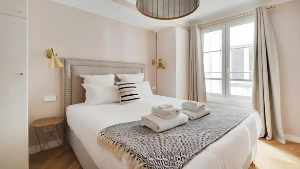 Una cama bien hecha con un plaid estampado, una lámpara de noche y una mesita redonda.