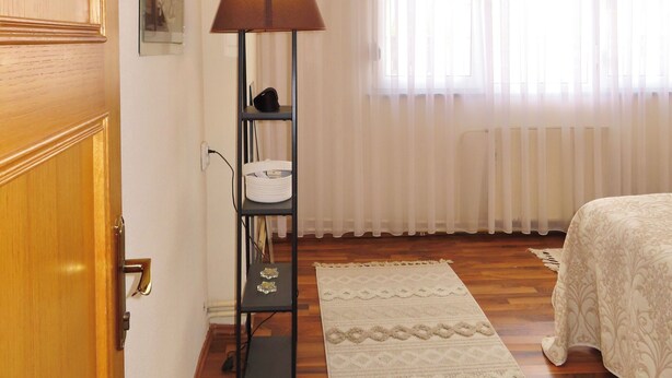 A hotel room with a wooden door, a standing lamp, a white rug, and a bed with a patterned bedspread. - Chez Apartment (Prizren)