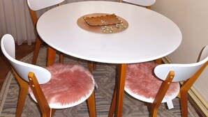 A round dining table with six white chairs, each with a wooden frame and a pink furry seat. A woven placemat is placed on the table. - Chez Apartment (Prizren)