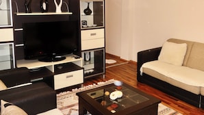 A modern living room with a flat-screen TV, dark sofas, a glass coffee table, and a decorative cabinet with glass doors. - Chez Apartment (Prizren)