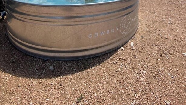 Una piscina metálica redonda con la marca "Cowboy", rodeada de grava, y un árbol, con un cielo azul despejado con nubes al fondo.
