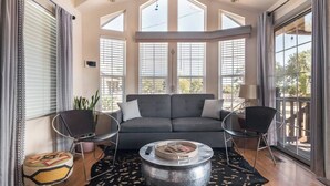 Een moderne woonkamer met een grijze bank, twee zwarte stoelen, een ronde salontafel en grote ramen met zonwering.