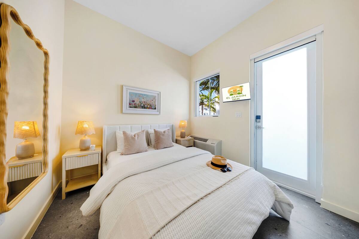 Un dormitorio con cama, lámparas de noche, un espejo, un cuadro en la pared y una ventana con vista a palmeras.