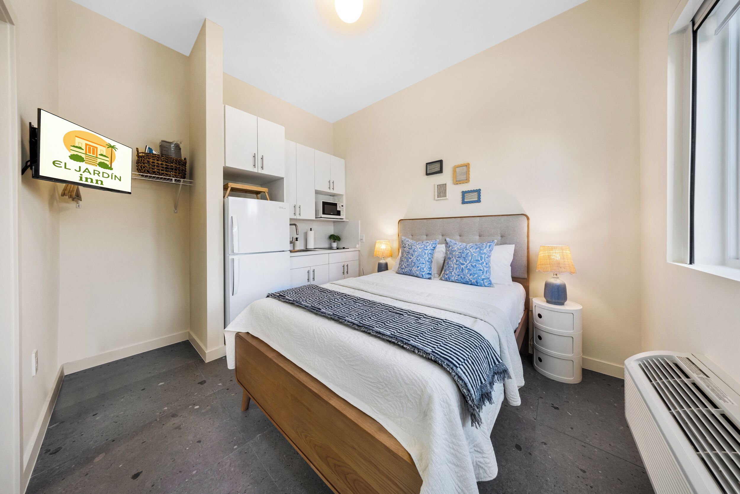 A bedroom with a bed, bedside tables, a nightstand, a wall-mounted TV, a kitchen area with cabinets and a microwave, and a window.