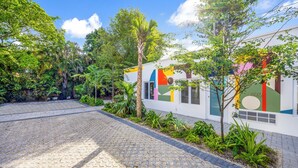 A paved courtyard with a white building featuring colorful geometric patterns, surrounded by lush greenery and trees. - El Jardin Inn (Miami)