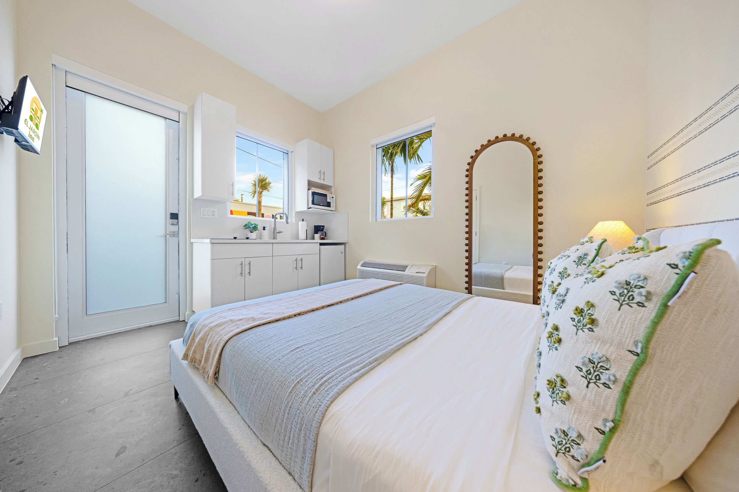 Une chambre à coucher comprenant un lit, un bureau, une chaise, un miroir et une fenêtre donnant sur des palmiers.
