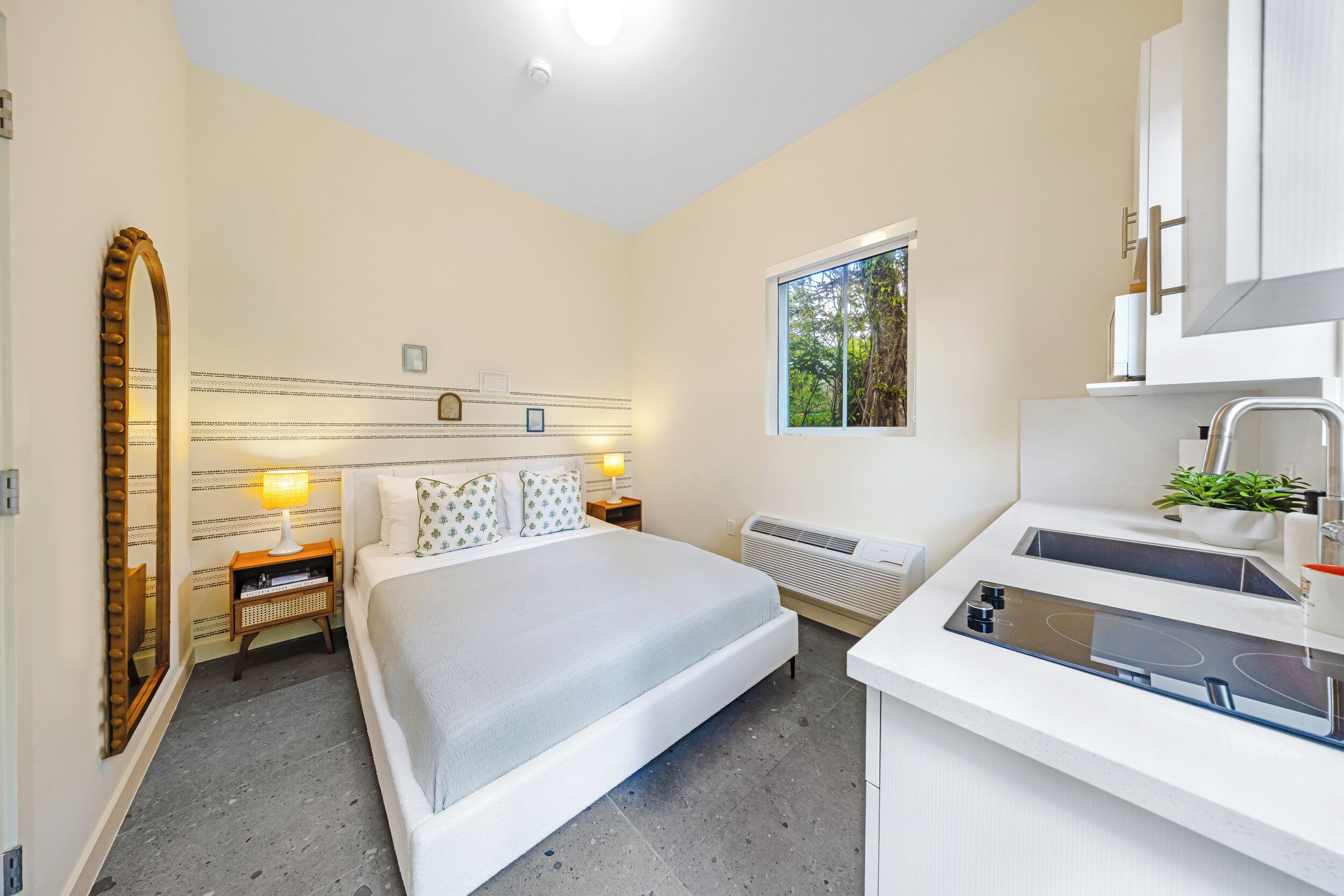 Ein modernes Hotelzimmer mit einem großen Bett, Nachttischlampen, einem kleinen Nachttisch, einem Fenster mit Aussicht, einer Küchenzeile mit Herd und einem Spiegel.