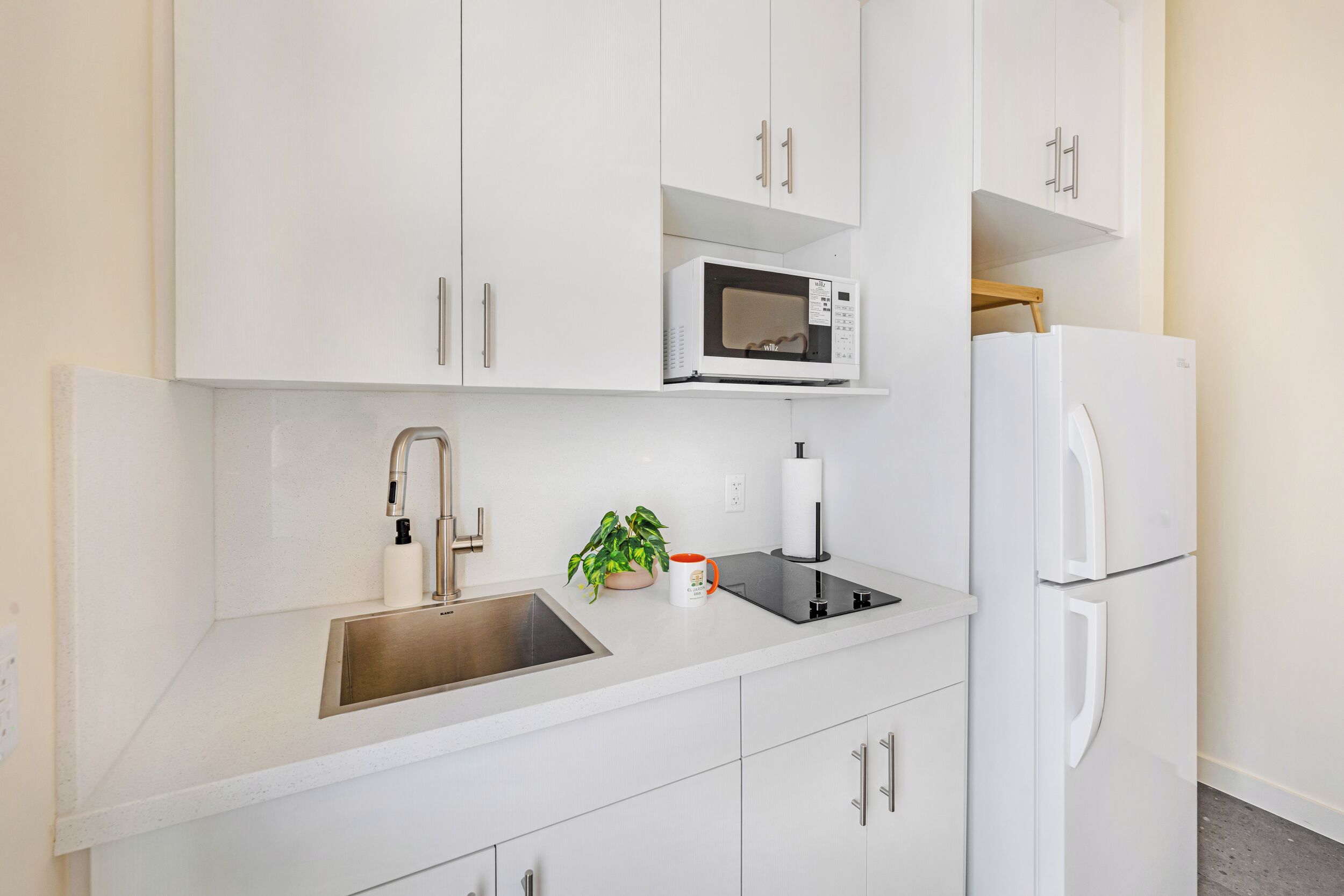A modern kitchen with white cabinetry, a stainless steel sink, a microwave, a refrigerator, and a small potted plant.