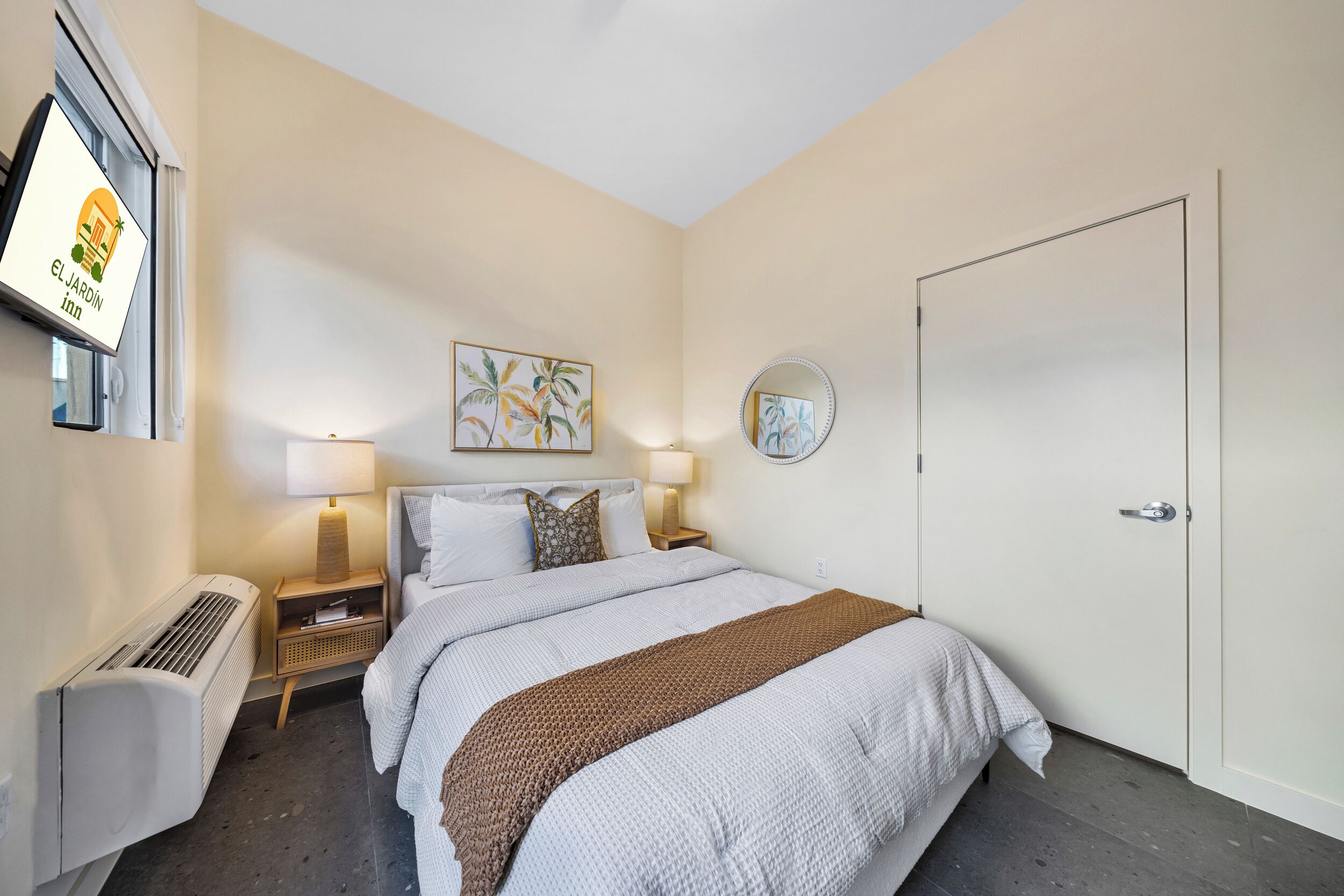 Habitación de hotel con cama, dos mesitas de noche con lámparas, un televisor montado en la pared, un cuadro, un espejo y una puerta.