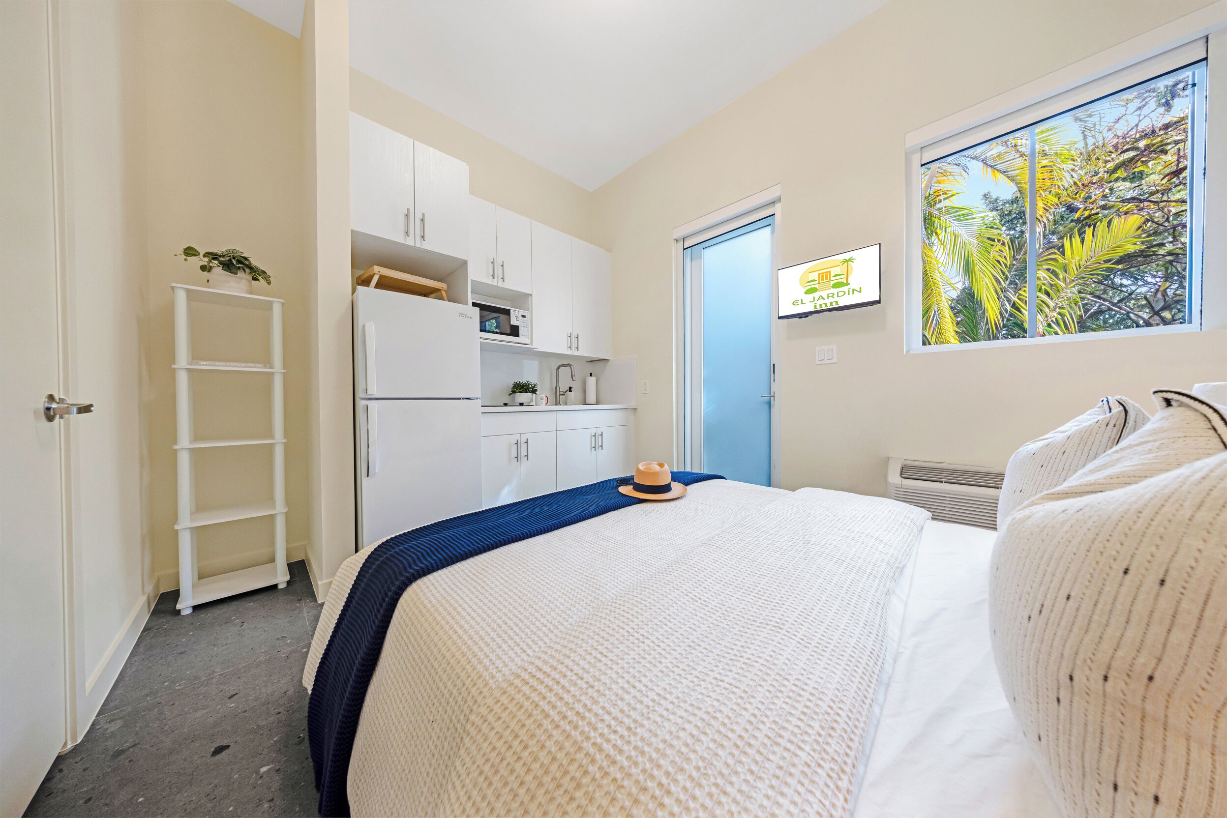 A bedroom with a bed, a wardrobe, a small kitchenette, a window with a view of palm trees, and a door to another room.