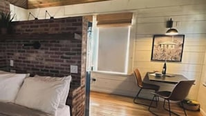 Un dormitorio con piso de madera, una cama con ropa de cama blanca, un escritorio con silla y una ventana con persianas.