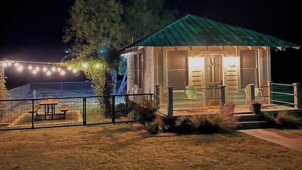 Un edificio pequeño de una sola planta con techo verde, un patio vallado con mesa de picnic y luces colgantes.