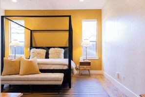 A bedroom with a four-poster bed, a bench, two bedside lamps, and a window with blinds.
