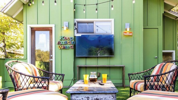 Uma casa verde com área de estar externa, uma TV fixada na parede e um aviso de 'Não Fumar'.