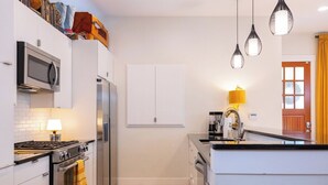 Cozinha compacta com armários brancos, micro-ondas, fogão e geladeira. O chão tem um padrão quadriculado, há um abajur amarelo e luminárias pendentes.