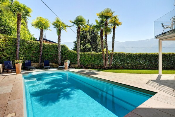 A swimming pool with palm trees, a hedge, and a view of mountains.