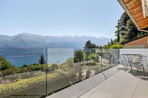 A terrace with a glass railing, outdoor seating, and a view of mountains and a lake.