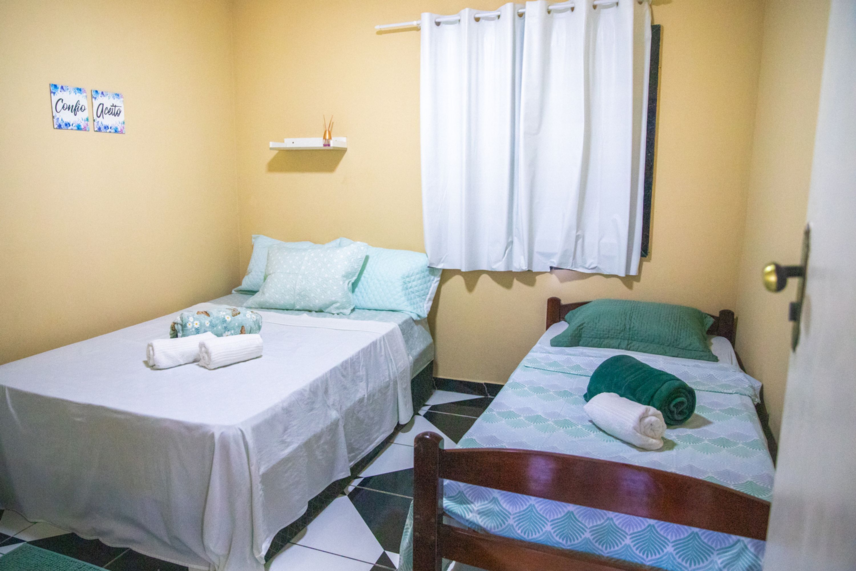 A small, clean room with two single beds, each with a pillow and a rolled towel. There is a shelf with a small item on it, and a window with white curtains.